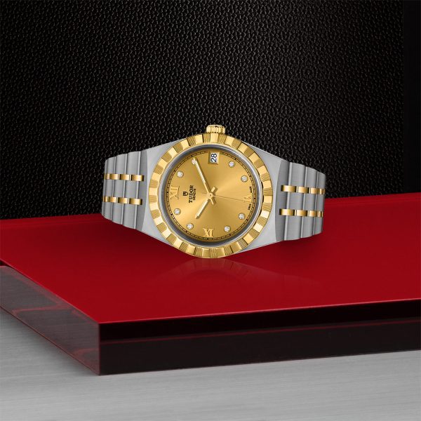 The TUDOR Royal, a luxury watch with a two-tone design featuring a gold dial, gold bezel, and a silver and gold bracelet, rests on a red surface. The timepiece displays the brand logo, has diamond hour markers, and includes a date window at the 3 o'clock position.