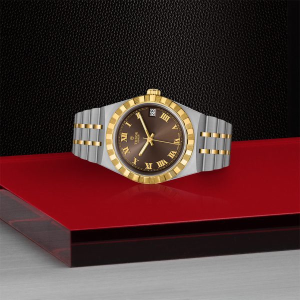 The TUDOR Royal, a luxury wristwatch featuring a black face adorned with gold Roman numerals and a striking gold and silver band, is showcased on a red and black surface. It boasts a fluted gold bezel and includes a date window at the 3 o'clock position.