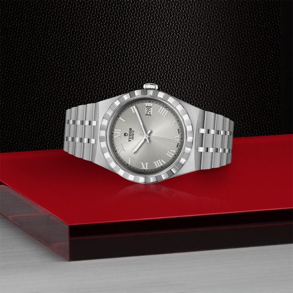 A silver TUDOR Royal watch with a Roman numeral dial and a metal link band is displayed on a red and grey surface against a black textured background. The watch features a date window at the 3 o'clock position.