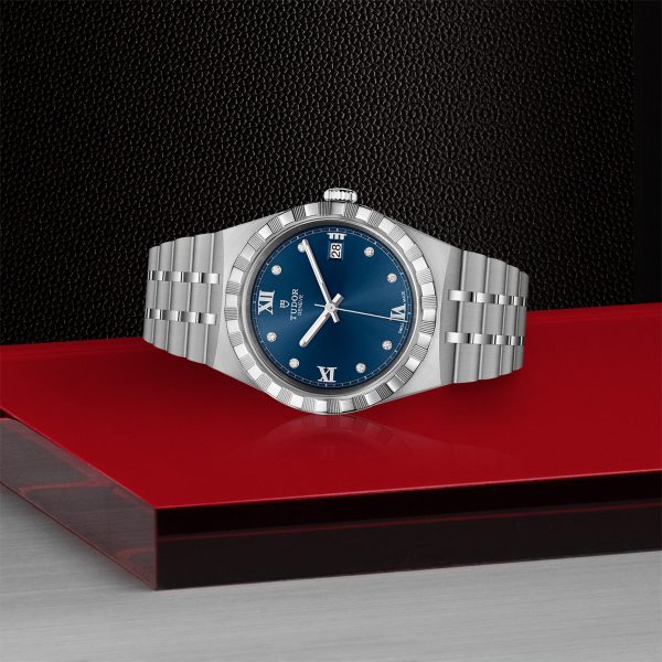 The TUDOR Royal wristwatch, featuring a blue dial and a silver stainless steel band, is showcased in a sleek, minimalist setting. This luxury timepiece boasts Roman numeral hour markers, a date window, and a fluted bezel, all elegantly presented on surfaces blending red, black, and gray tones.