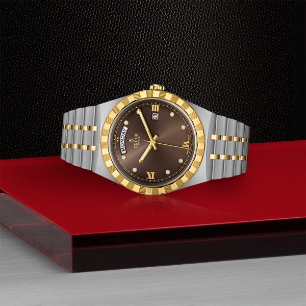 The TUDOR Royal wristwatch with a silver and gold bracelet is elegantly displayed on a red and black platform. This luxury watch boasts a brown dial, gold Roman numerals, and a day-date function positioned at the top and right side. The watch is encased in a gold and silver bezel and case.