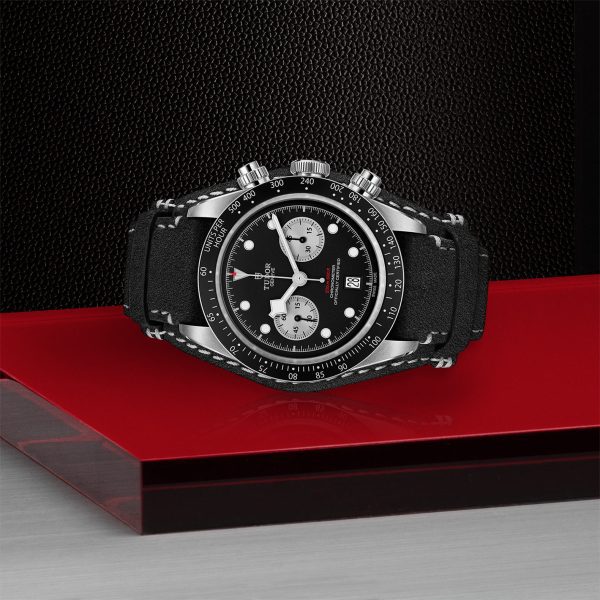 The TUDOR Black Bay Chrono, showcasing a black dial with chronograph sub-dials and a matching black strap, is displayed. This wristwatch features elegant silver accents, luminous hands, and markers and is set against a textured black and red background.