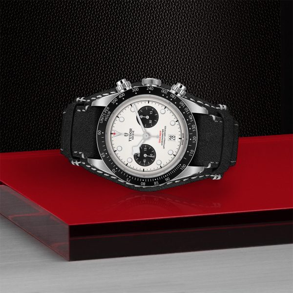 The TUDOR Black Bay Chrono, a black and silver chronograph wristwatch with a white face, is displayed on a red and gray pedestal. The watch features two sub-dials, a tachymeter bezel, and a black leather strap. The background boasts a textured black surface.