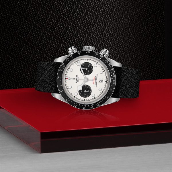 The TUDOR Black Bay Chrono is showcased, highlighting its luxury design with a white dial and black subdials. The watch features a striking black bezel with white markings and comes with a sophisticated black textured strap. The background includes textured black surfaces complemented by smooth red and gray elements.