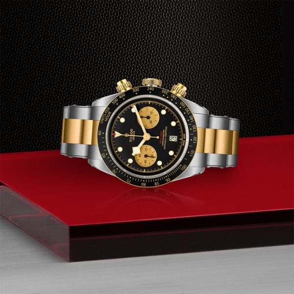 The TUDOR Black Bay Chrono S&G 41mm Steel and Gold wristwatch, featuring a sophisticated black dial, gold accents on the face and bezel, and a chic two-tone metal strap in gold and silver, is showcased against a striking red, black, and gray background. This luxurious timepiece also includes two subdials and a date window.