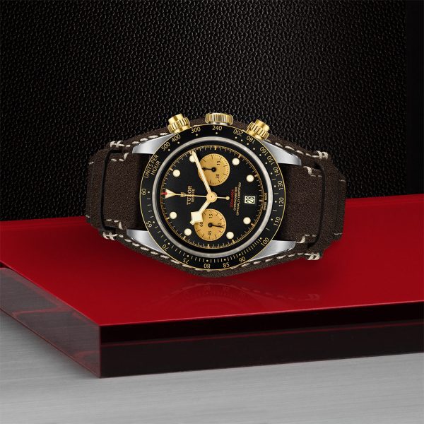 The TUDOR Black Bay Chrono S&G 41mm Steel and Gold wristwatch features a black dial with gold subdials and a laid-back brown leather strap, showcased on a stylish red and black display stand.