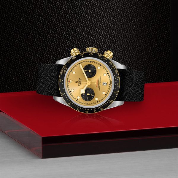 The TUDOR Black Bay Chrono S&G, a luxury wristwatch with a black textured strap, is displayed on a red and gray platform. The watch face features a gold dial with three black subdials, gold hour markers, and two gold pushers on the side, all set against a black tachymeter bezel. The background is textured black.