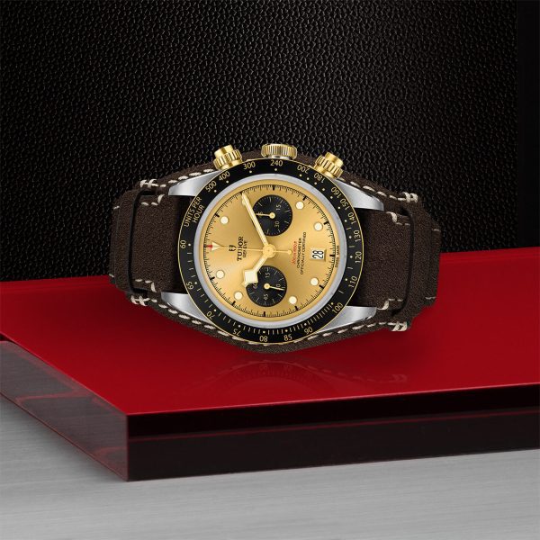 The TUDOR Black Bay Chrono S&G, featuring a striking black and gold face, is elegantly displayed. Its golden bezel, two black sub-dials, date window, and sophisticated black and brown leather strap make a bold statement against a background of black, red, and grey that highlights the watch’s luxurious design.