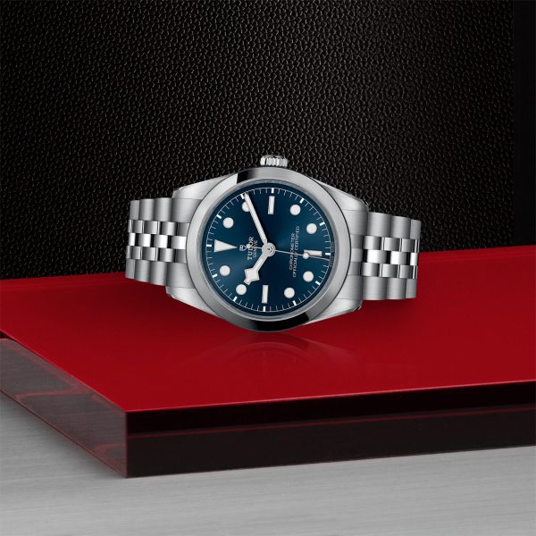 The TUDOR Black Bay 36, a luxury wristwatch in silver with a blue dial and a metal strap, is showcased on a red and black platform against a dark textured background. The watch face features hour markers and simple, elegant hands.