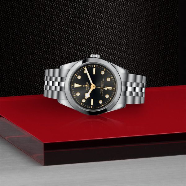 The TUDOR Black Bay 36, a luxury wristwatch with a black dial and gold accents, is placed on a red surface. The watch features a stainless steel bracelet with alternating polished and brushed links. The background consists of a black textured surface.