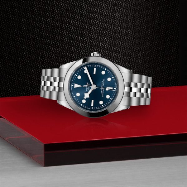 The TUDOR Black Bay 39 wristwatch, featuring a dark blue dial with silver markers, is elegantly displayed on a red surface against a textured black background. The watch showcases a stainless steel bracelet and boasts a classic design with a round face.