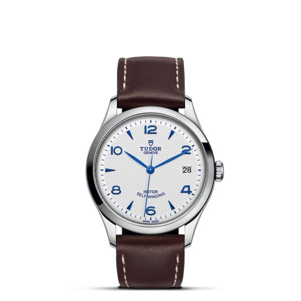 The TUDOR 1926 wristwatch features a white dial with blue Arabic numerals and hands. It includes a date window at the 3 o'clock position, the Tudor logo at 12 o'clock, and "ROTOR SELF-WINDING" inscribed at 6 o'clock. The watch is paired with a brown leather strap accented by white stitching.