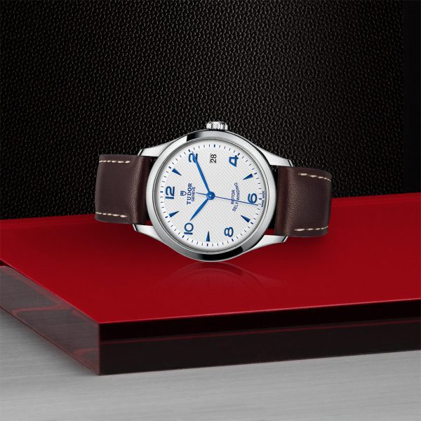 The TUDOR 1926 luxury wristwatch, featuring a brown leather strap, silver casing, and white dial with blue numerals and hands, is displayed on a red surface against a black textured background. The brand name "Tudor" and "Geneva" are clearly visible on the dial.