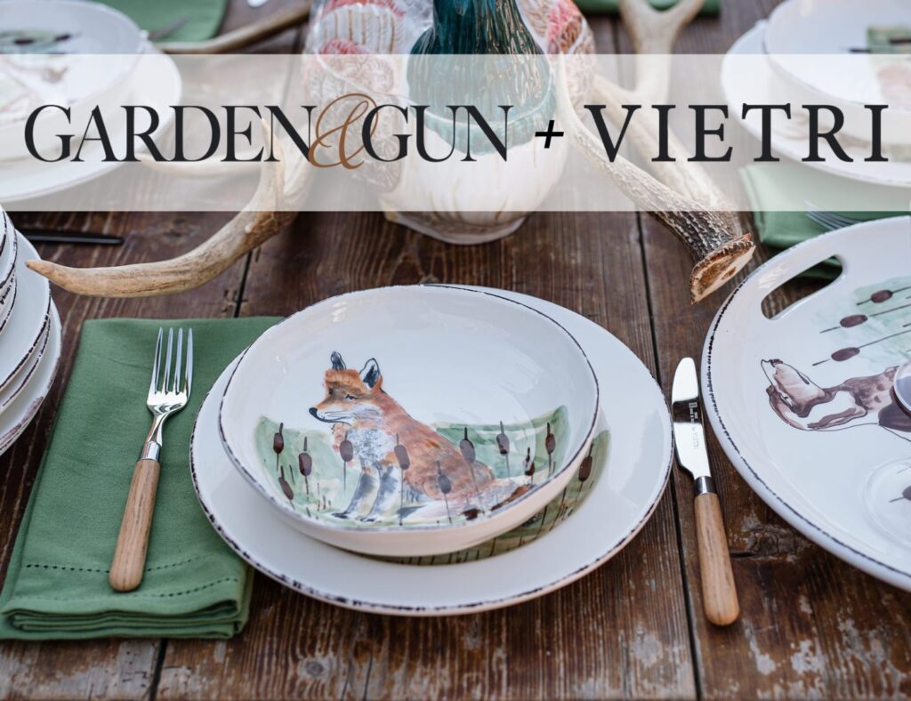 A rustic wooden table is set with plates, bowls, and cutlery. The main plate design features a fox illustration. Napkins are green. The table also has decorative pumpkins and antler accents. Overlaid text reads "GARDEN & GUN + VIETRI.