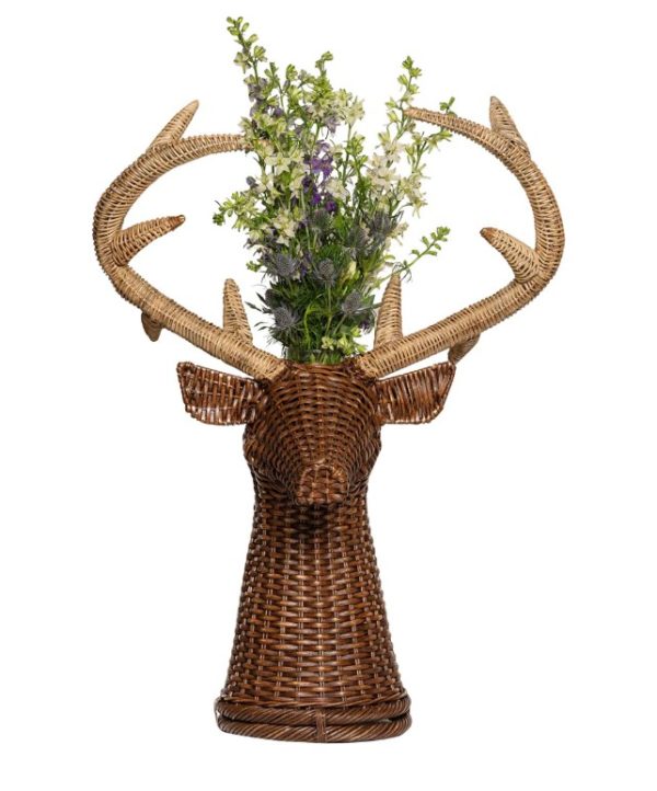 The Juliska Devon Willow 31" Stag Vase, designed as a wicker sculpture of a deer's head with large antlers, functions as a unique vase holding a vibrant arrangement of greenery along with purple and white flowers. The earthy tones and natural materials imbue it with rustic, artistic appeal.