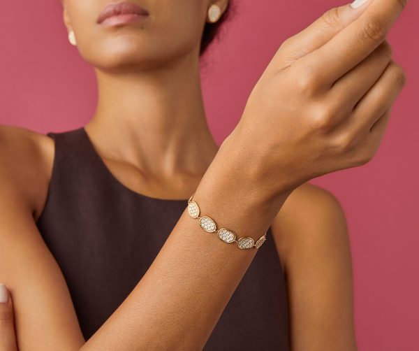 A person poses against a pink background, wearing a sleeveless brown top. They have their arm raised to showcase the Marco Bicego Lunaria Pave Diamond Link Bracelet, which features oval, textured links in gold.