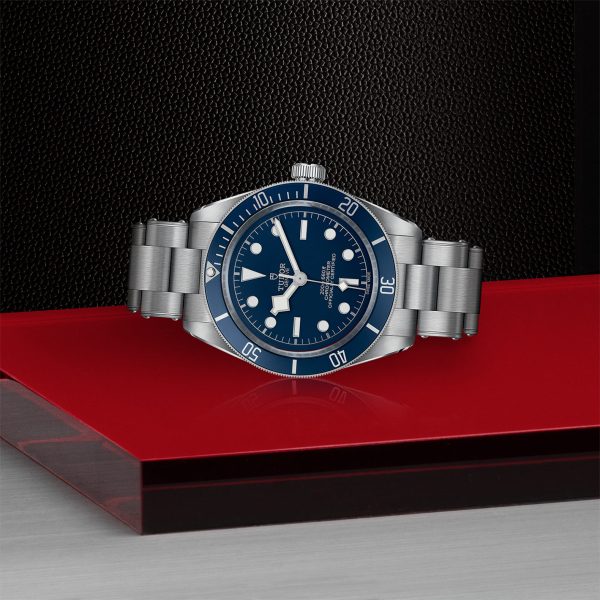 The TUDOR Black Bay Fifty-Eight, a stainless steel wristwatch with a blue bezel and dial featuring luminescent hour markers, is displayed on a red and black surface with a textured background. The watch boasts a metal link bracelet and prominent white hands.