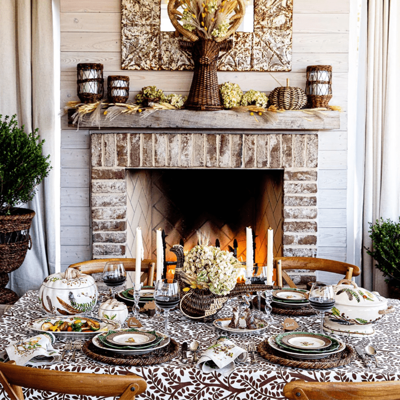 A cozy dining room with a brick fireplace and a beautifully set tablescape for a meal. The table features a patterned tablecloth, plates, wine glasses, candles, and a centerpiece with flowers. Rustic decor, including wicker vases and natural elements, adorns the mantel.