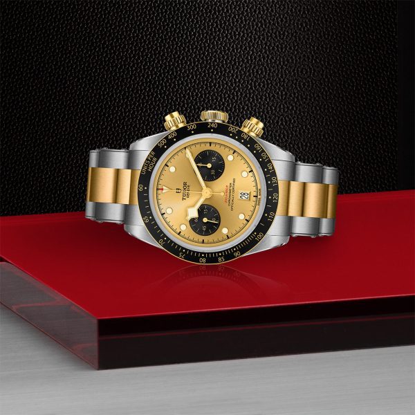 The TUDOR Black Bay Chrono S&G wristwatch, boasting a gold and silver metal band, is showcased against a textured black backdrop with contrasting red and gray surfaces. This luxury timepiece features a gold face accented by black subdials, a date window, and a tachymeter bezel.