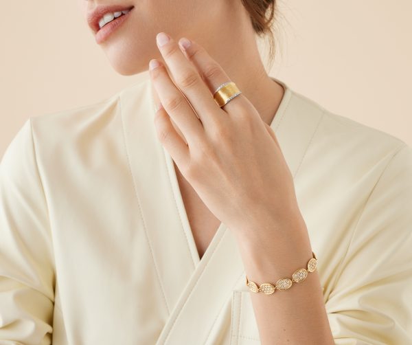 A person clad in a cream-colored outfit is touching their chin, showcasing the Marco Bicego Lunaria Diamond Band on their finger and a gold bracelet featuring oval links adorned with small crystals. The background is soft and neutral in tone.