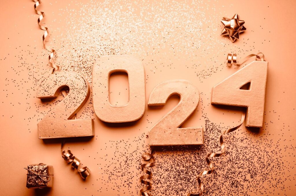 Golden "2024" numerals surrounded by sparkling glitter, golden ribbon curls, a small wrapped gift, and a decorative bow, all set against a matching golden backdrop celebrating the New Year with the Pantone Color of the Year.