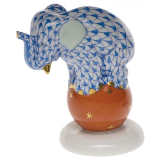 The Herend Dancing Elephant Figurine - Blue features a porcelain elephant shaped figurine decorated with an intricate blue and white scale pattern, standing on its hind legs atop a round, orange base adorned with stars. The figurine is positioned on a smooth, white platform.