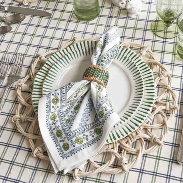 A table setting showcasing the Juliska Sitio Stripe Dessert/Salad Plate in Basil on a wicker placemat. A white napkin adorned with green and blue floral patterns is secured by a beaded napkin ring. The tablecloth features a green and blue plaid pattern, complemented by green glasses and silver cutlery.