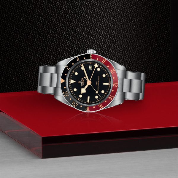 The TUDOR Black Bay 58 GMT is showcased on a glossy red surface, featuring a stainless steel band, a black dial, and a striking two-tone bezel in red and black. The luminescent hour markers and hands further enhance its sophisticated design. A textured black background accentuates the watch's elegance.