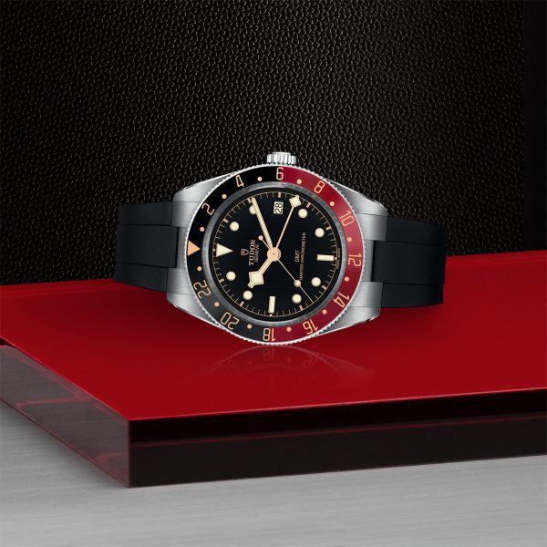 A TUDOR Black Bay 58 GMT wristwatch sits on a red and black display. The watch features a stainless steel case, black dial, and dual-color bezel in red and blue. It is fitted with a black rubber strap and has luminous hour markers as well as a date window.