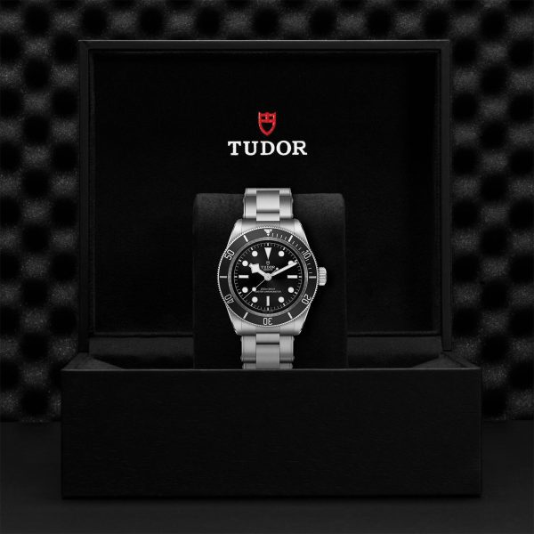 A TUDOR Black Bay wristwatch with a silver case, black dial, and matching black bezel is displayed inside an open black box. The Tudor logo and name are prominently shown on the inside lid of the box, which has a textured background. The watch is centered and positioned upright on a black cushion.