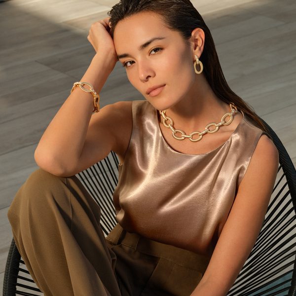 A woman with long, dark hair is seated, resting her head on one hand. She's wearing a bronze-colored satin sleeveless top, brown pants, and statement jewelry, including hoop earrings and a matching bracelet. Around her neck is the Roberto Coin Duchessa Diamond Oval Link Collar that complements her thoughtful gaze at the camera.