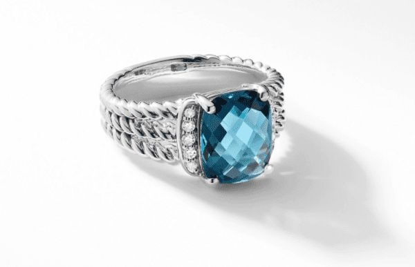 The PETITE WHEATON RING IB boasts a large, faceted blue gemstone at its center, complemented by small diamonds on either side. The silver band highlights a twisted rope design that adds texture and elegance. Displayed on a white, reflective surface, the ring exudes sophistication.