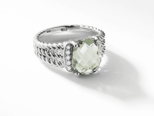 The PETITE WHEATON RING PL is a silver ring adorned with a prominent, faceted, translucent gemstone at its center, complemented by small round diamonds on either side. The band boasts an intricate rope-like design that adds both texture and elegance to the piece. The ring is elegantly displayed against a white background.