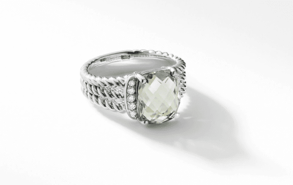 A close-up image of the PETITE WHEATON RING PL showcasing a large, faceted clear gemstone at the center. The silver band features an intricate braided design, adorned with small, round diamonds flanking the central gemstone on both sides. The ring is set against a white background.