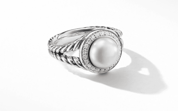 The 8MM Petite Cerise Ring PE is a stunning silver piece, showcasing a central round pearl encircled by a halo of delicate clear gemstones. Its band features an intricate twisted rope design, adding to its elegant and sophisticated charm. The ring casts a gentle shadow on the white background.