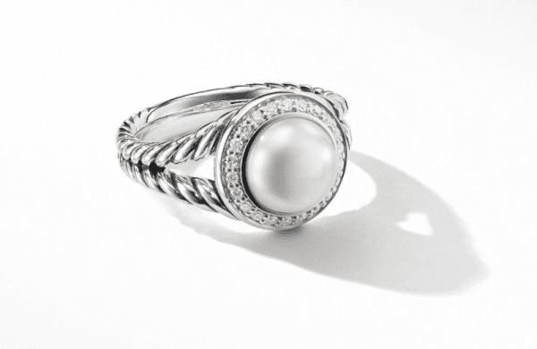 The 8MM Petite Cerise Ring PE showcases a prominent central pearl, encircled by a band of small diamonds. The twisted, rope-like design of the silver band adds an elegant touch. The ring is displayed against a plain, white background with a shadow cast beside it.