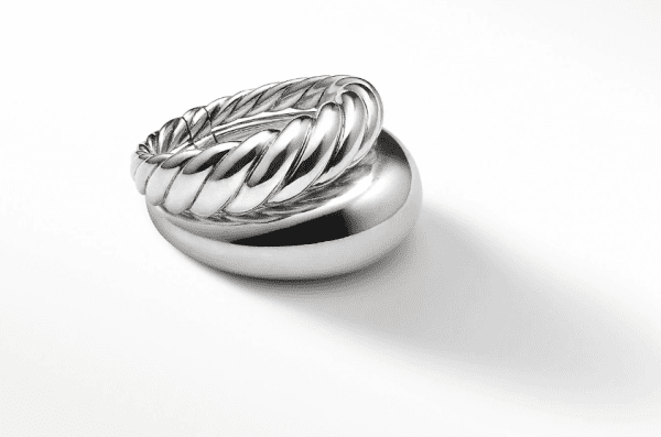 The PURE FORM 2 ROW RING, a shiny twisted silver ring with a ribbed design, is elegantly displayed on a sleek, reflective silver oval base against a plain white background. The ring casts a soft shadow to the right.