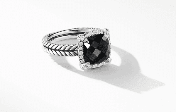 The 9MM CHATELAINE PAVE BEZEL RING BO features a black faceted gemstone set in the center, encircled by a border of small clear stones. The silver band boasts a twisted rope design, casting a shadow on the white background.