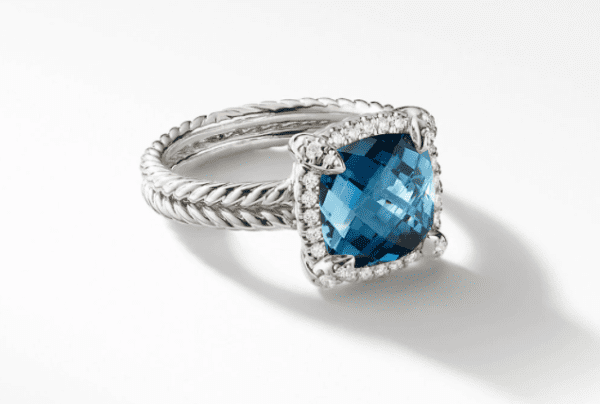 The 9MM Chatelaine Pave Bezel Ring IB features a large, square-cut blue gemstone at its center, surrounded by small clear stones. The band showcases an intricate twisted rope design. The ring is displayed on a white background, casting a subtle shadow.