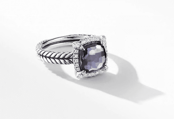 The 9MM CHATELAINE PAVE BEZEL RING AH boasts a twisted silver band, showcasing a square-shaped, faceted purple gemstone centerpiece encircled by small clear stones. The ring is elegantly displayed on a white surface, casting a soft shadow.