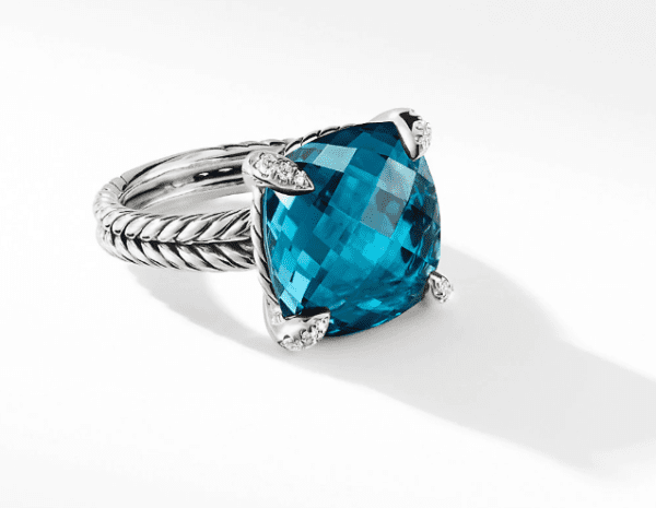 The 14MM CHATELAINE RING BT features a sophisticated silver twisted band design, with a substantial square-cut blue gemstone at its center, secured by four prongs. Each prong is embellished with tiny, brilliant diamonds that accentuate the ring's detailed and elegant look. The ring gently casts a soft shadow.