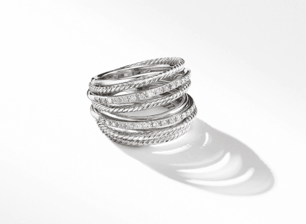 The CROSSOVER WIDE RING, a silver accessory featuring multiple intertwined bands with varying textures such as smooth and rope-like patterns, and adorned with small sparkling stones, is displayed on a white background. Shadows from the ring's intricate design are visible on the surface.