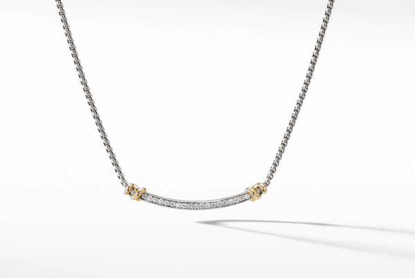 A close-up view of the HELENA DIAMOND STATION NECKLACE showcases its thin chain, featuring a curved bar pendant encrusted with small diamonds and accented by delicate gold knots on either side. The necklace is displayed against a white background.