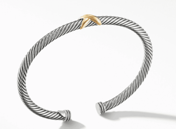 The 4MM X BRACELET is a sleek, twisted cable bracelet crafted from silver, showcasing an open cuff design. It features a small, gold, X-shaped accent prominently positioned at its top center. Displayed against a clean white surface with a subtle shadow beneath it.