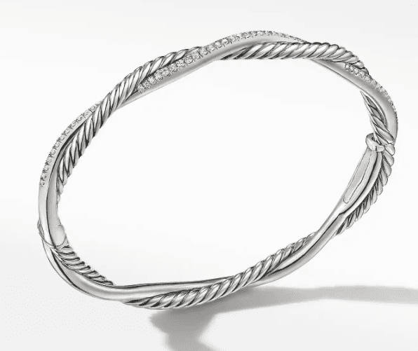 The PETITE INFINITY BRACELET is a sleek, silver-toned twisted bangle featuring a combination of smooth and braided textures with small sparkling stones embedded along parts of the design, set against a plain white background.