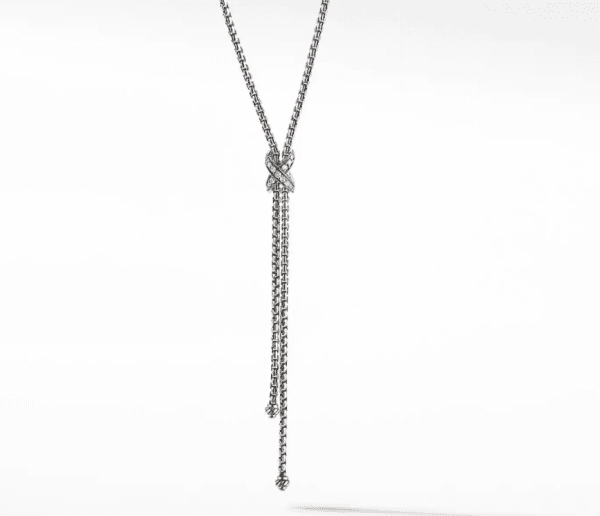 The PETITE X LARIAT is a silver lariat necklace showcasing a double-chain design with a central knot detail and two dangling chain ends. This necklace exudes elegance with its sleek and sophisticated appearance, set against a plain white background.