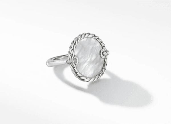 The 18MM ELEMENTS RING features an oval-shaped, pearl-like center stone encased in a twisted rope-style border, flanked by two small, round decorative elements. The ring casts a delicate shadow on a white background.