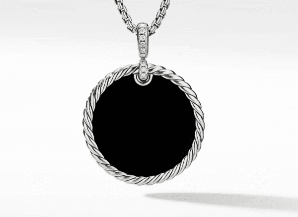 A close-up image of the 32MM REVERSIBLE ELEMENTS pendant hanging from a chain. The pendant showcases a sleek black surface, encircled by an elegant twisted silver border. The loop attaching the pendant to the chain is embellished with tiny, sparkling stones. The background is plain white.