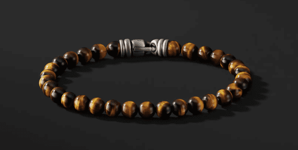 The 6MM SPIRITUAL BEAD bracelet showcases round tiger's eye gemstones that have a striking brown and black striped pattern. These beads are elegantly strung together with a sleek silver clasp and displayed against a dark background.