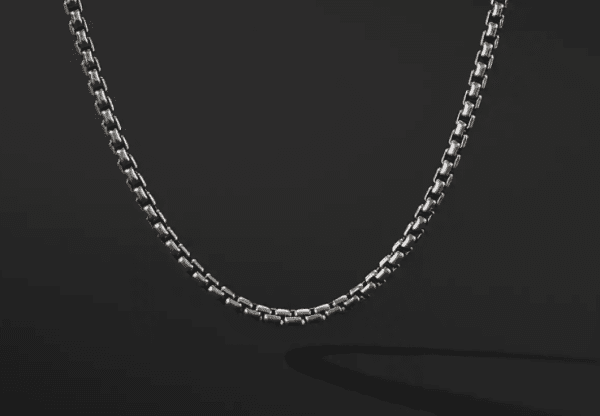 The 2.7MM MENS BOX CHAIN is showcased against a solid black background. The chain forms a gentle U-shape, highlighting its intricate links and smooth, metallic finish. Its subtle shadow appears beneath it on the dark surface.